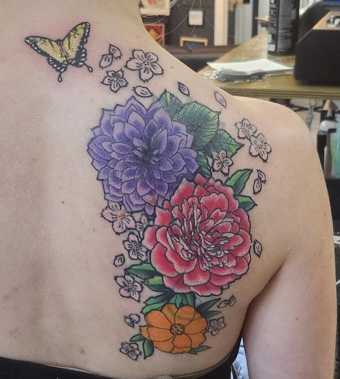 A colorful floral tattoo on a person's shoulder and upper back, featuring a purple dahlia, a red peony, an orange flower, and a yellow butterfly, with green leaves surrounding the flowers.