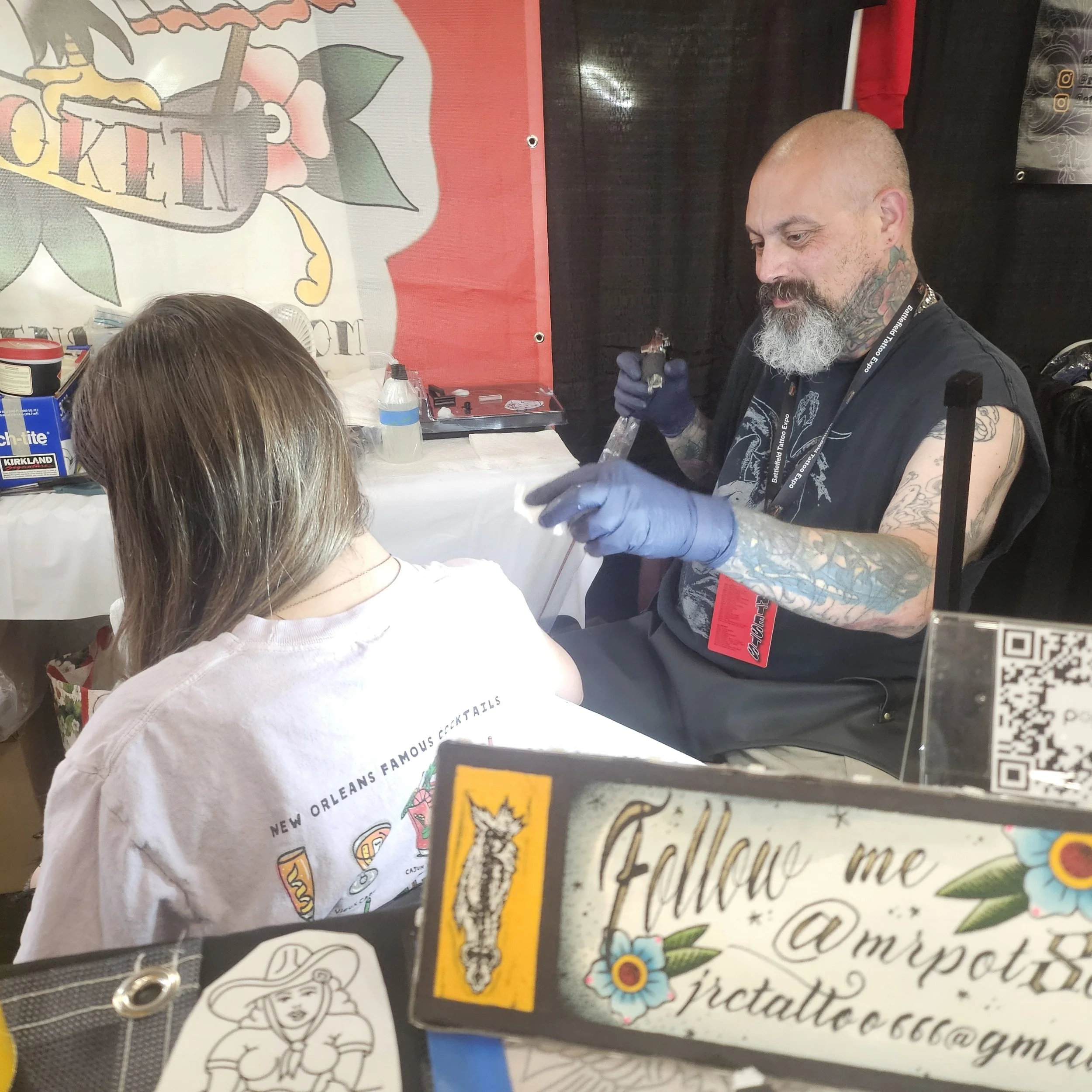 Tattoo artist with tattooed arms, wearing a black sleeveless shirt and blue gloves, is tattooing a woman with light brown hair and a white T-shirt, seated at a table with tattoo supplies, in an indoor tattoo studio.