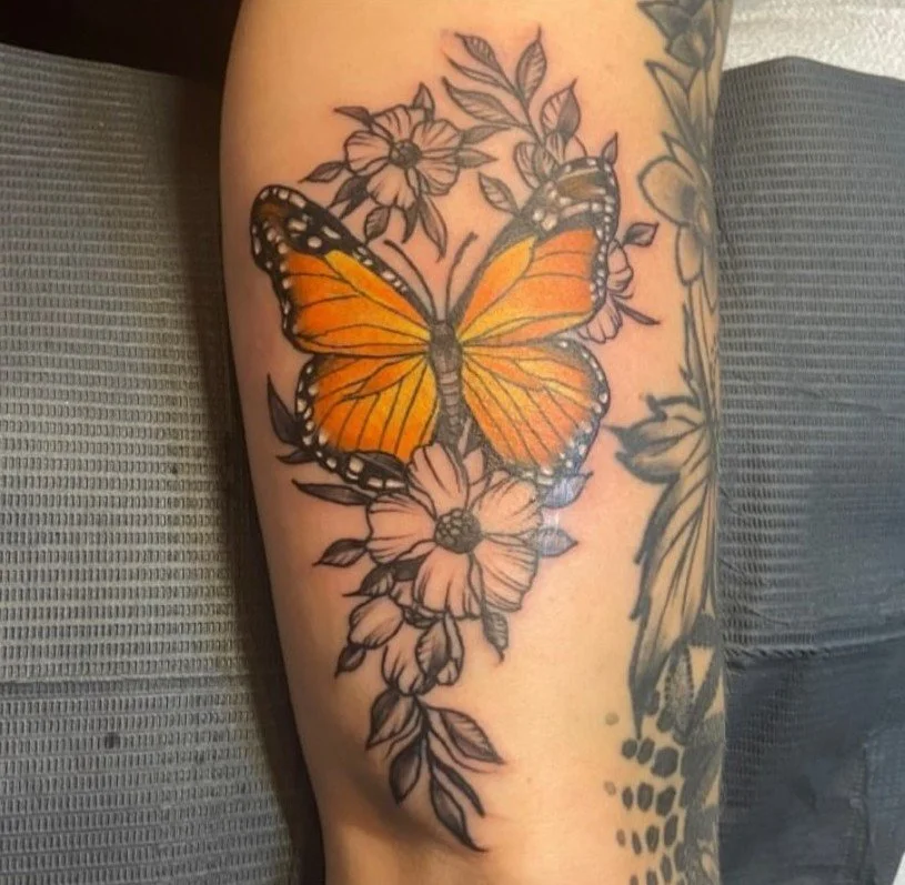 A tattoo of an orange butterfly with black and white markings, surrounded by black-and-white floral designs, on a person's arm.