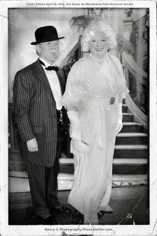 A black-and-white photo of a man in a suit with a bow tie and a woman in a dress with a large bow on her head, standing together and smiling.