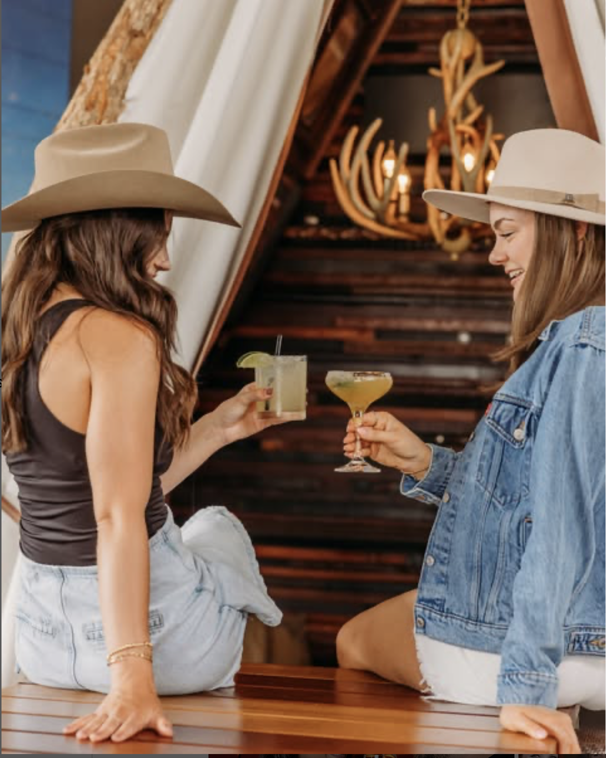 Two women wearing cowboy hats sitting on a wooden bar, each holding a cocktail; one drinks a yellow beverage with a lime wedge, the other a greenish drink with a straw; warm lighting and rustic background
