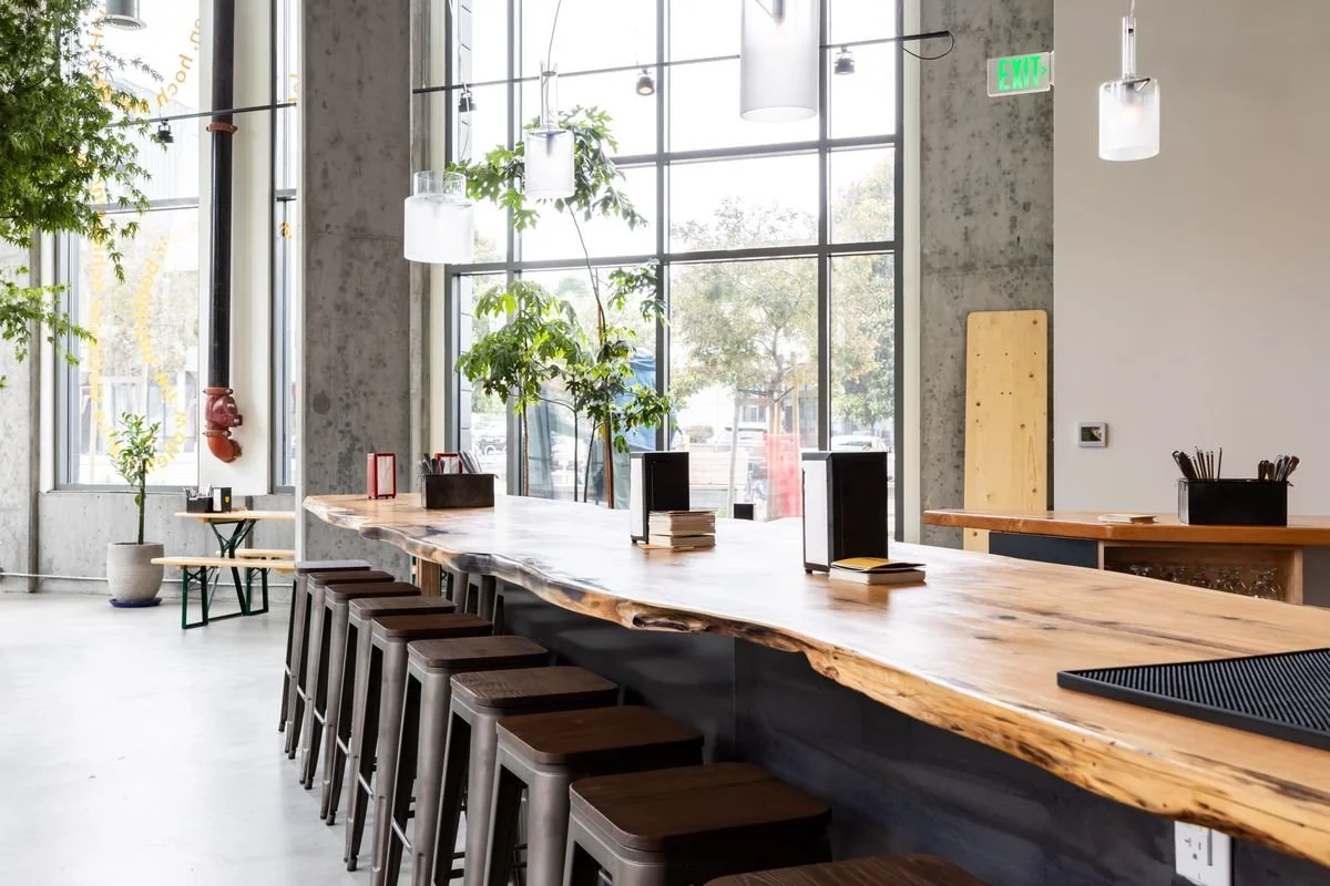 A modern, minimalist cafe interior with a large wooden bar counter, wooden stools lined up, potted plants, large windows, and pendant lights.