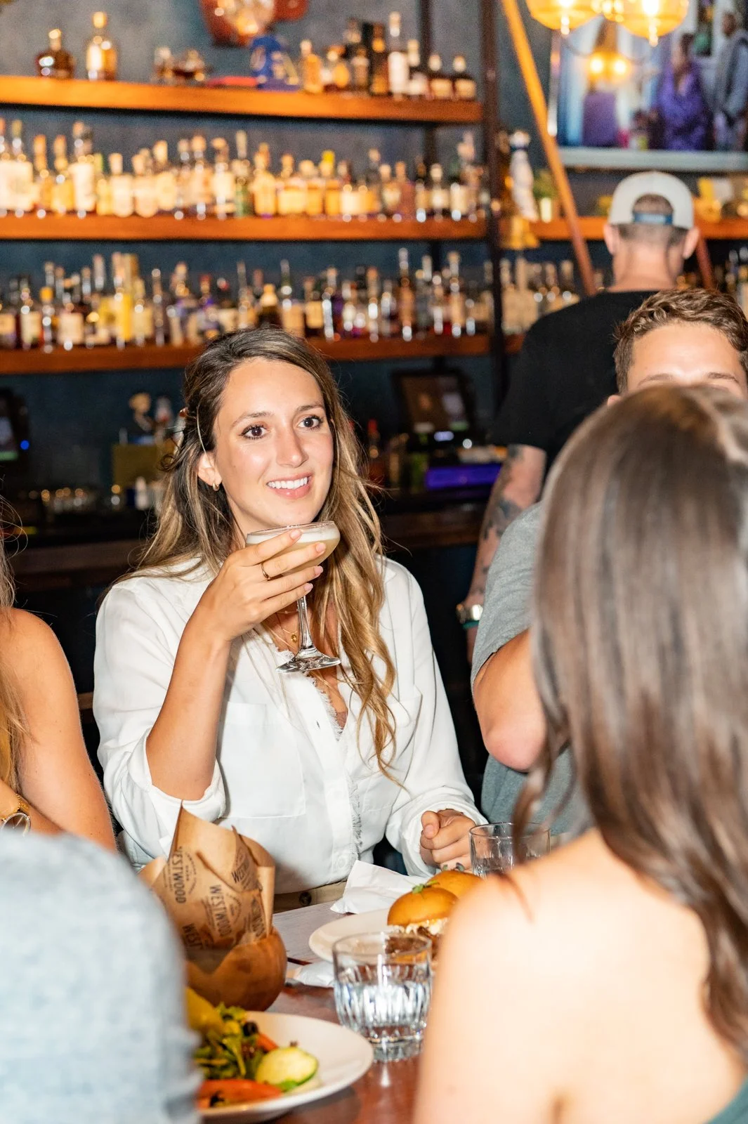 A woman with long wavy hair wearing a white shirt holding a cocktail at a social gathering in a bar or restaurant.