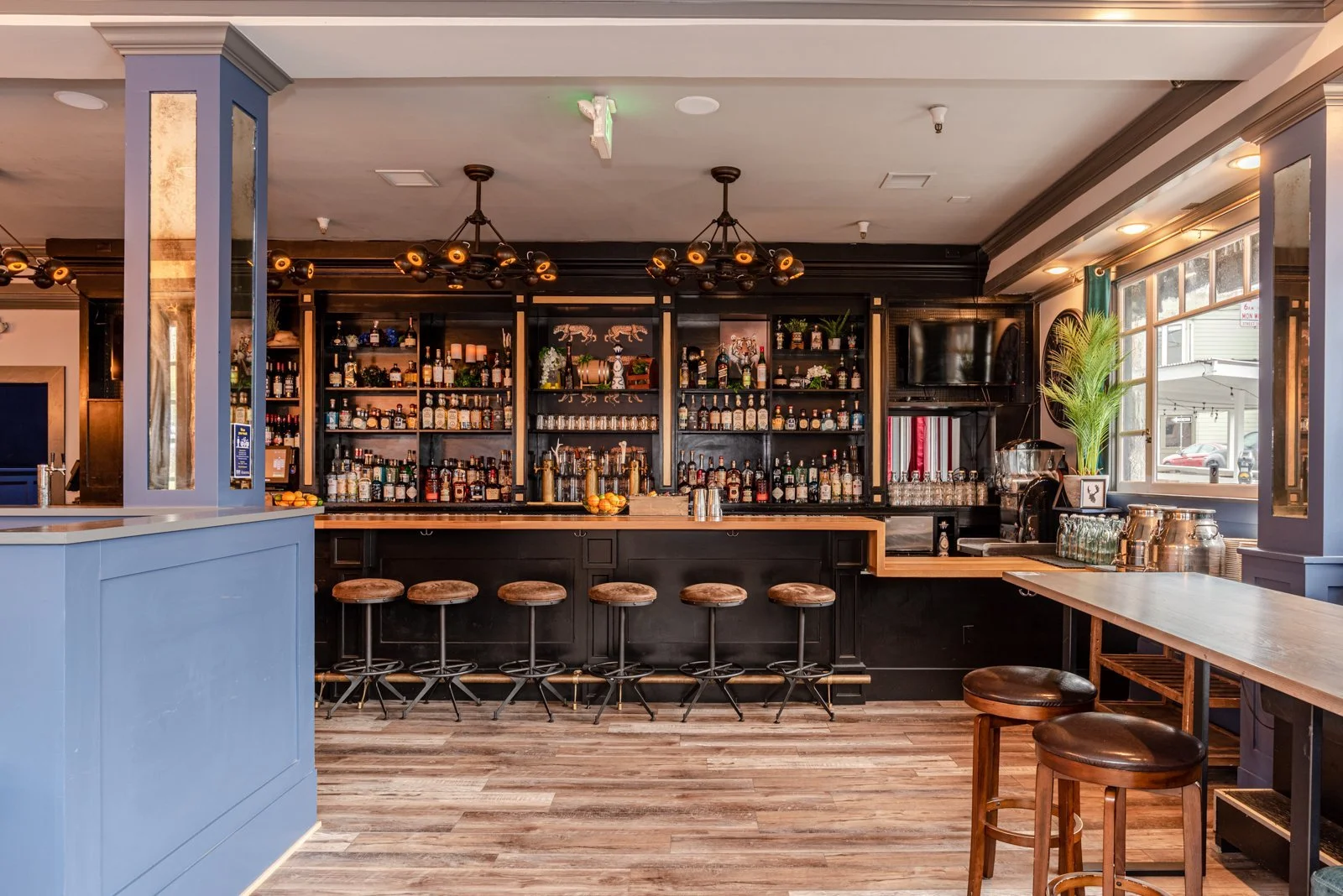 Bar interior with wooden bar counter, shelves stocked with liquor bottles, barstools, and a window with sunlight.
