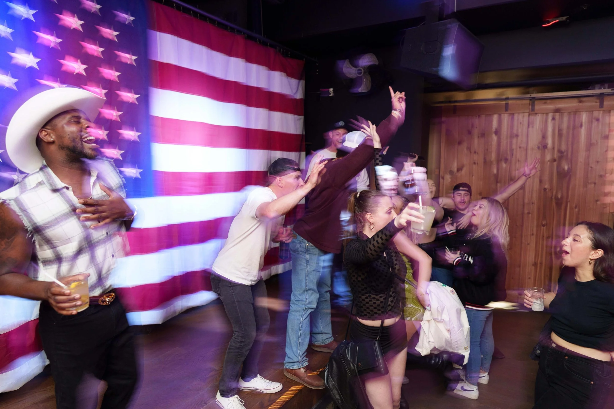 People dancing and enjoying drinks at a celebration with an American flag backdrop.