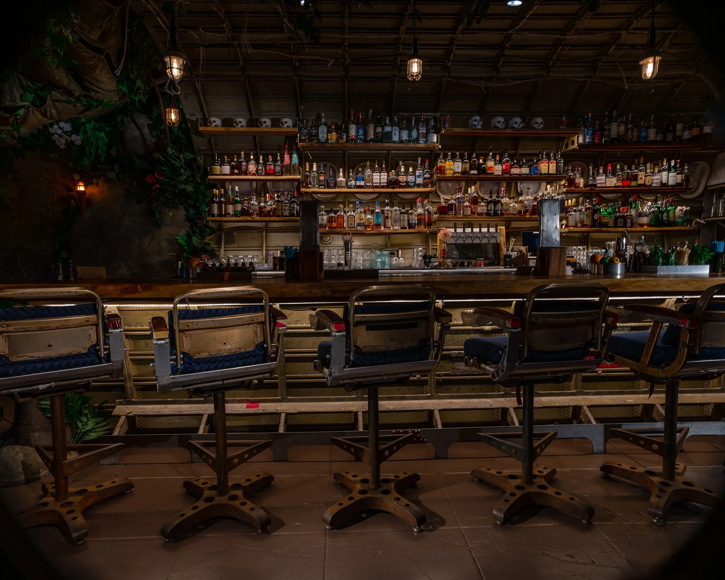 A dimly lit bar with high chairs in front of a long wooden counter. Behind the counter are shelves stocked with various bottles of alcohol and skull-shaped decorations on top. The bar has warm, ambient lighting and a cozy, rustic atmosphere.