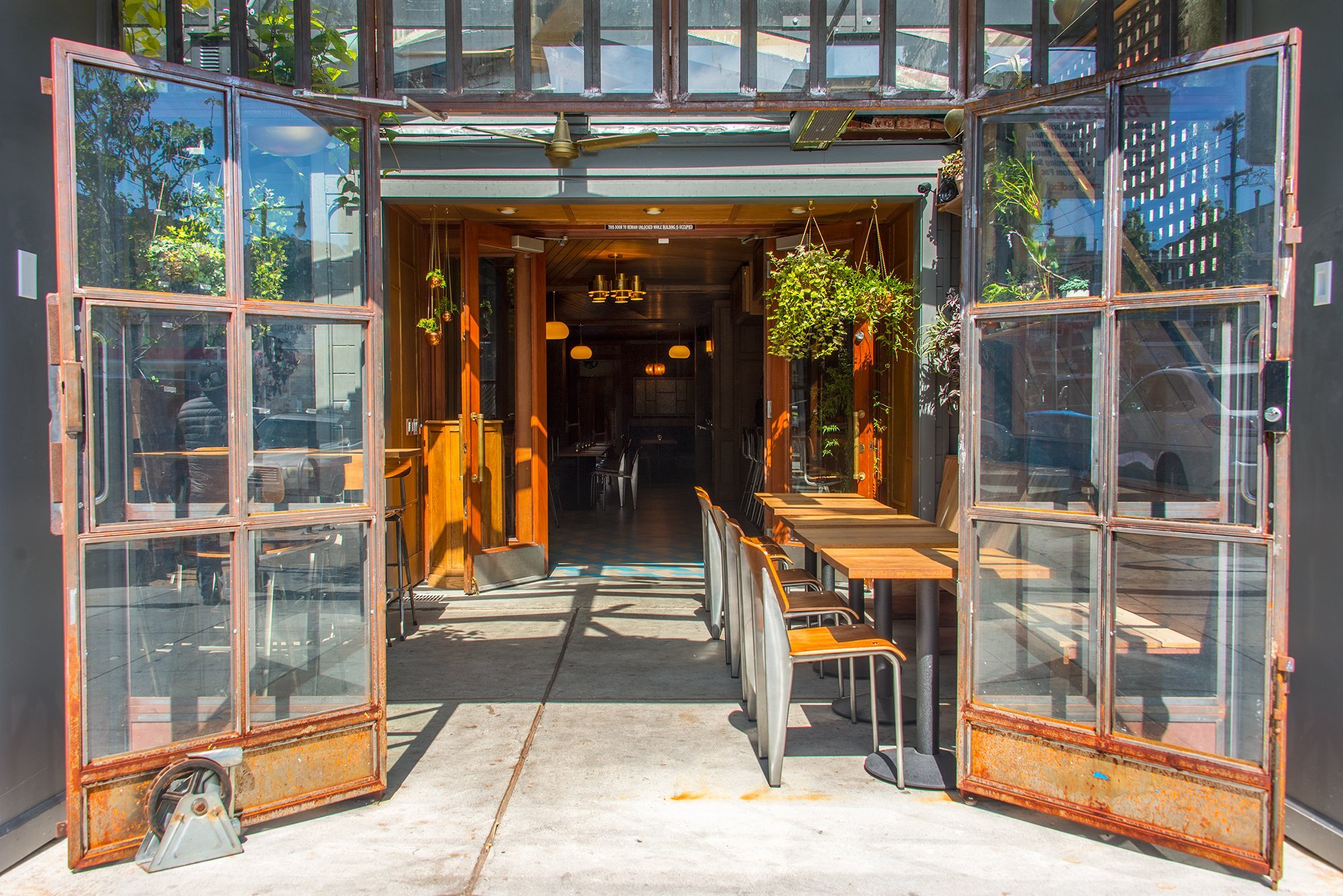 Open glass doors leading into a modern restaurant with wooden accents and hanging plants, illuminated by hanging light fixtures inside.