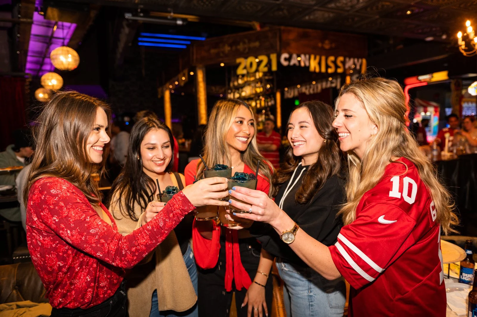 A group of six women celebrating with drinks at a bar, wearing casual clothes, one in a red sports jersey, in a lively decorated bar with a sign that reads '2021 CAN KISS MY' in the background.