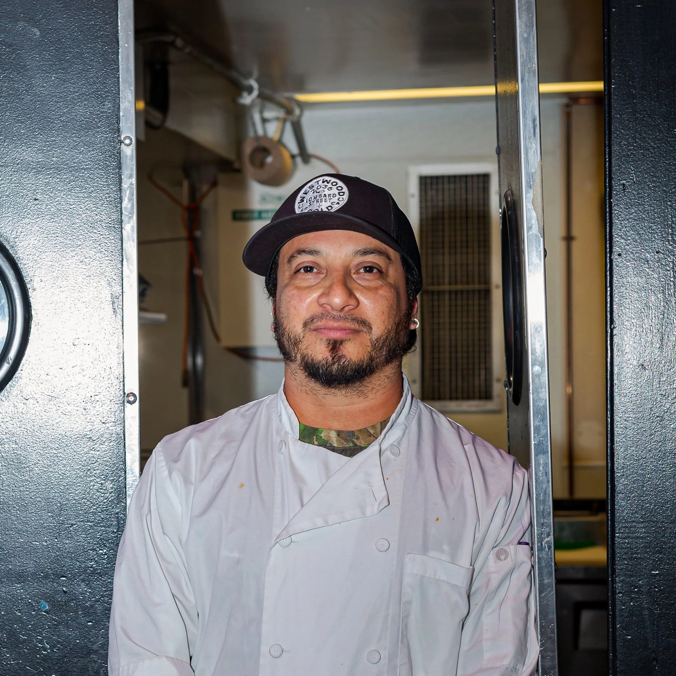 A man in a white chef's coat and black baseball cap standing in a kitchen doorway.