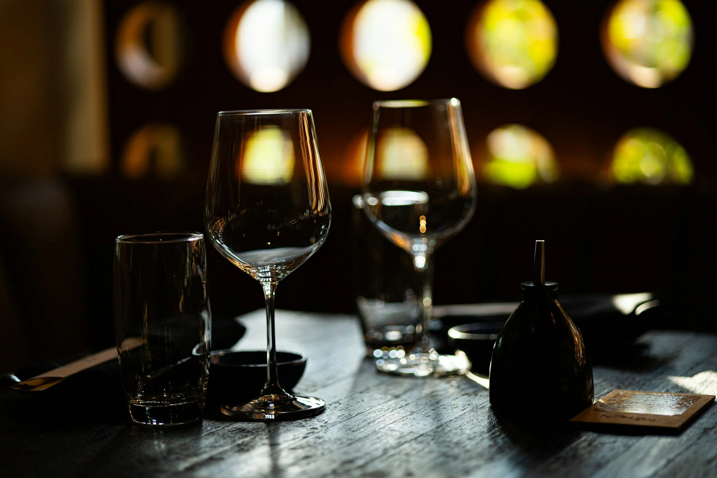 Empty wine glasses and drinking glass on a dark wooden table, with sunlight filtering through circular windows in the background.