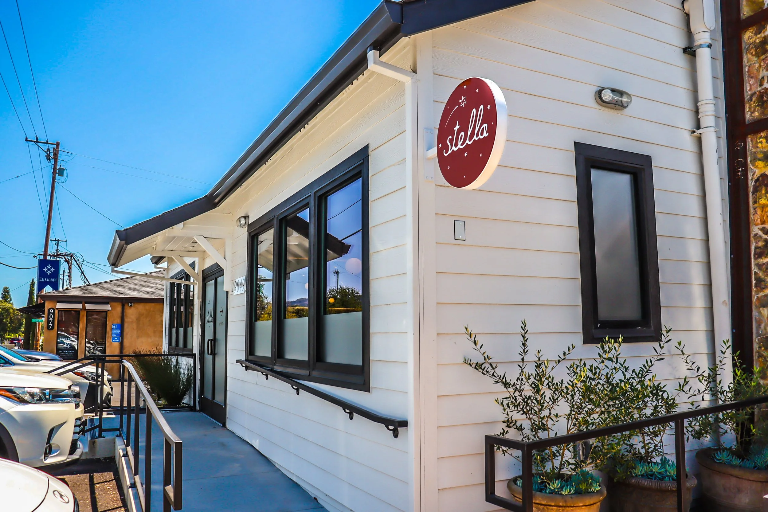 White building with a red circular sign that says 'stella', large windows, and a parking lot in front.