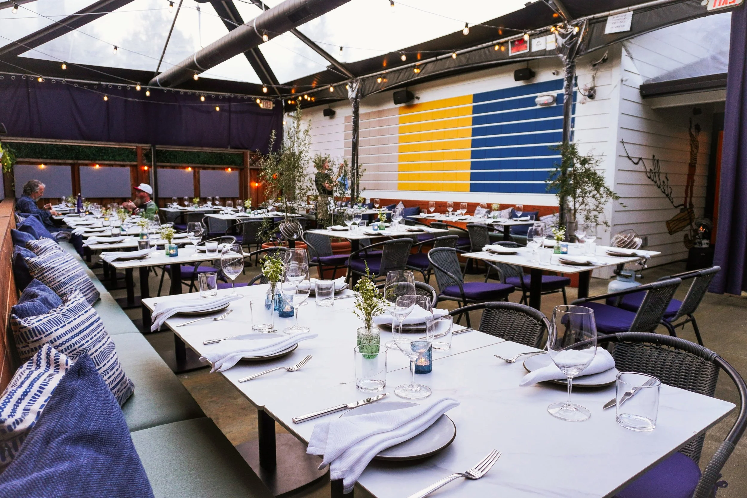 Decorated restaurant patio with white tables set with plates, glasses, napkins, and small flower arrangements. There are purple cushions on the bench seating and black chairs with purple cushions around the tables. The roof is transparent with string lights hanging. The background features a mural with yellow and blue stripes and a white wooden wall with mural art.