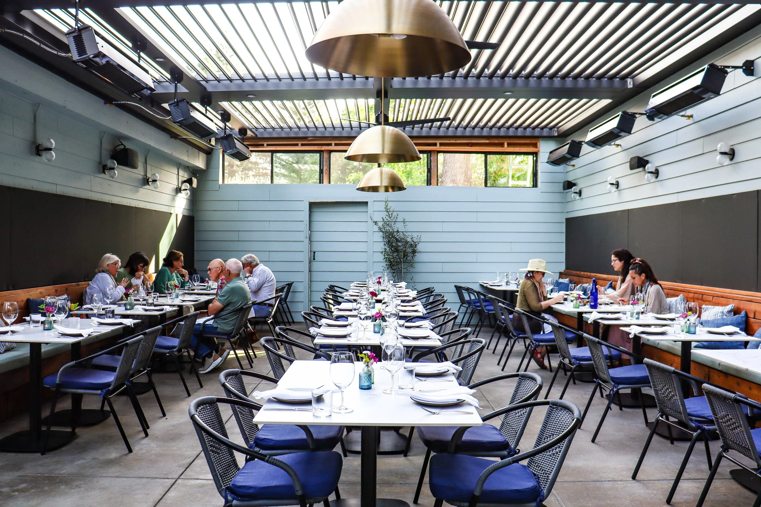 Interior of a modern restaurant with tables set with wine glasses, cutlery, and flowers, and people dining, with natural light coming through large windows and a ceiling with metal slats.