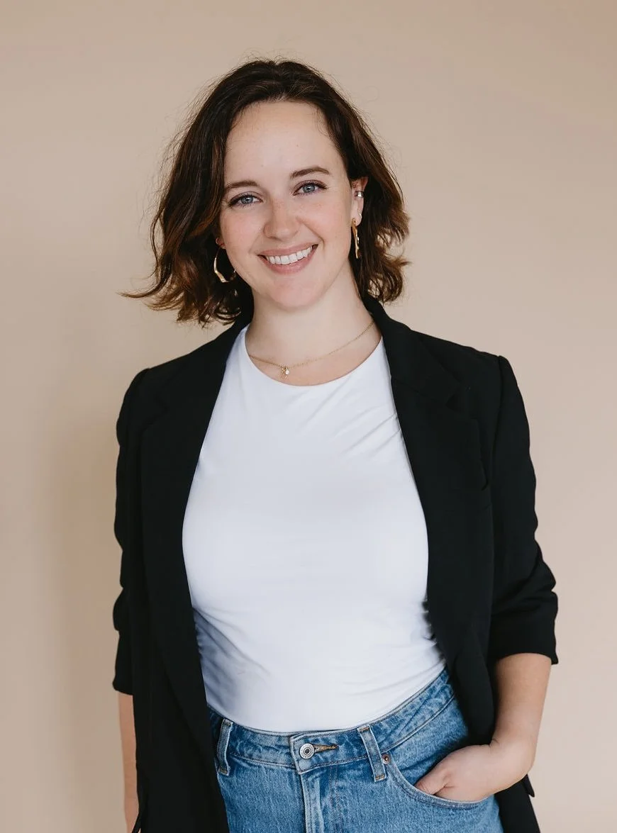 A young woman, Chelsea Kern, with shoulder-length brown hair and blue eyes, smiling, wearing a black blazer, white t-shirt, and blue jeans, standing in front of a beige background.
