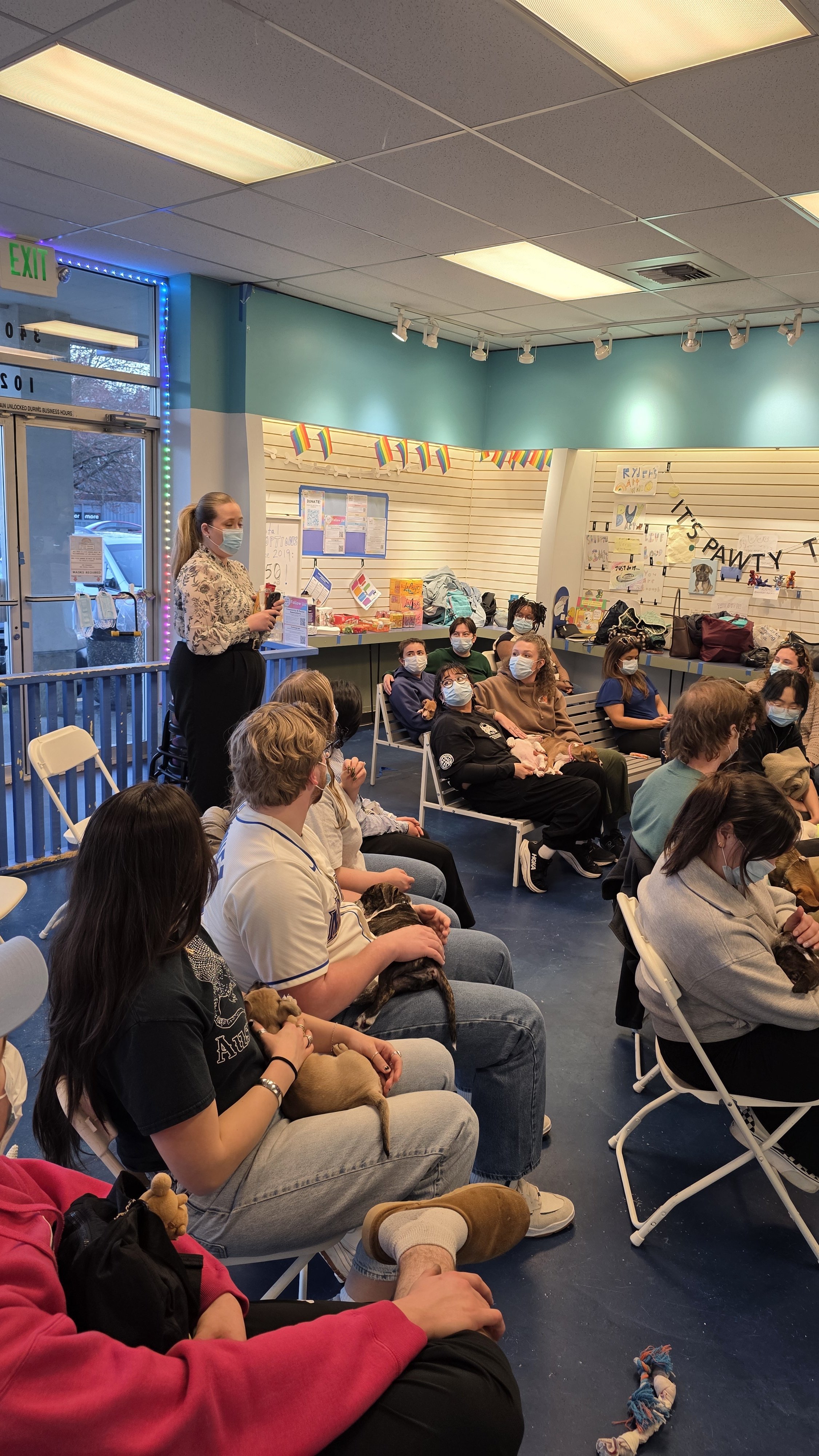 A group of people sitting and listening to a woman standing at the front in a room decorated with colorful banners and posters. Many attendees are wearing masks and some are holding dogs.