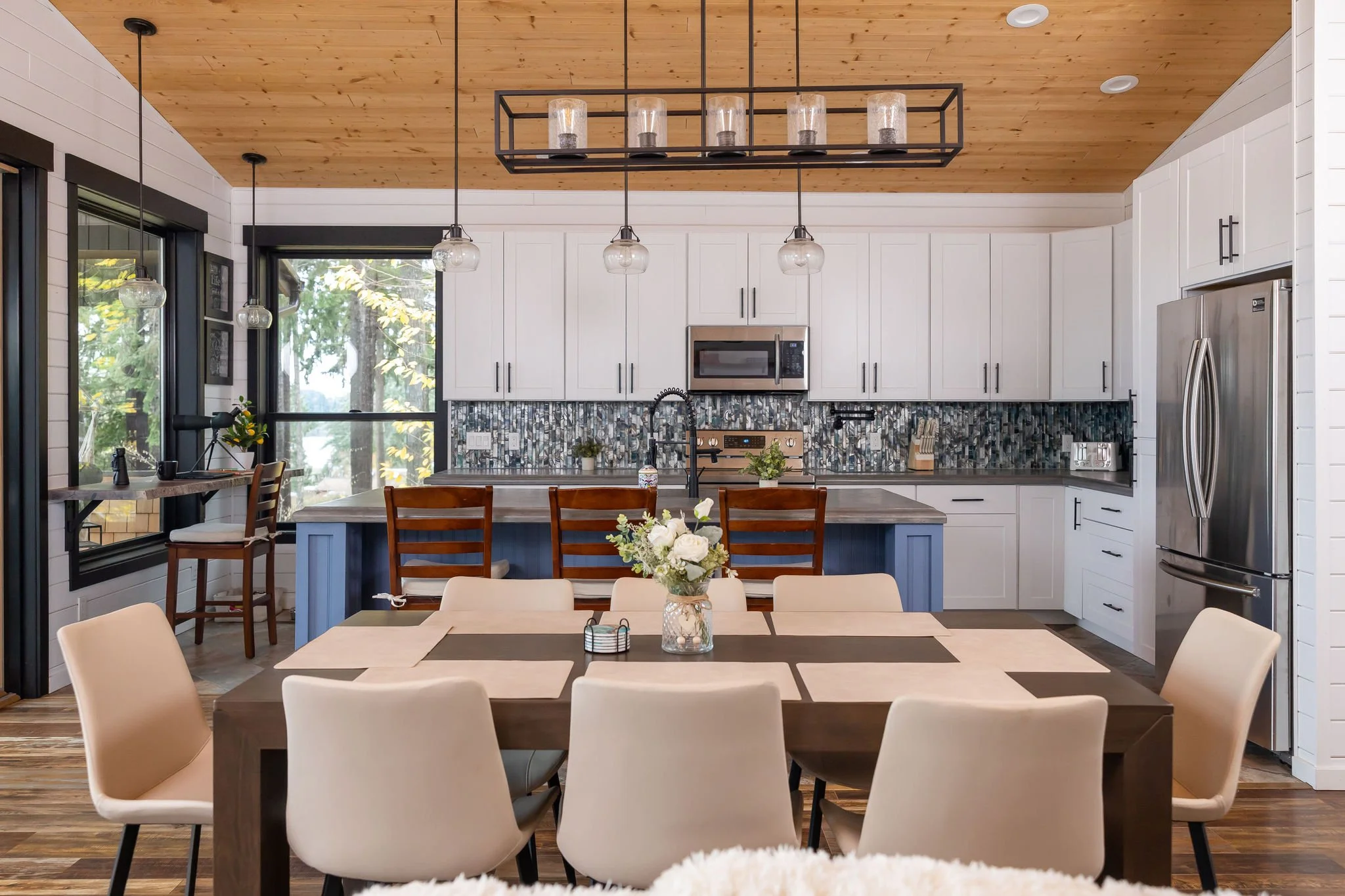 Modern kitchen with white cabinets, wooden ceiling, black hardware, stainless steel appliances, a mosaic tile backsplash, a kitchen island with seating, and a dining table with 8 beige chairs and a floral centerpiece.