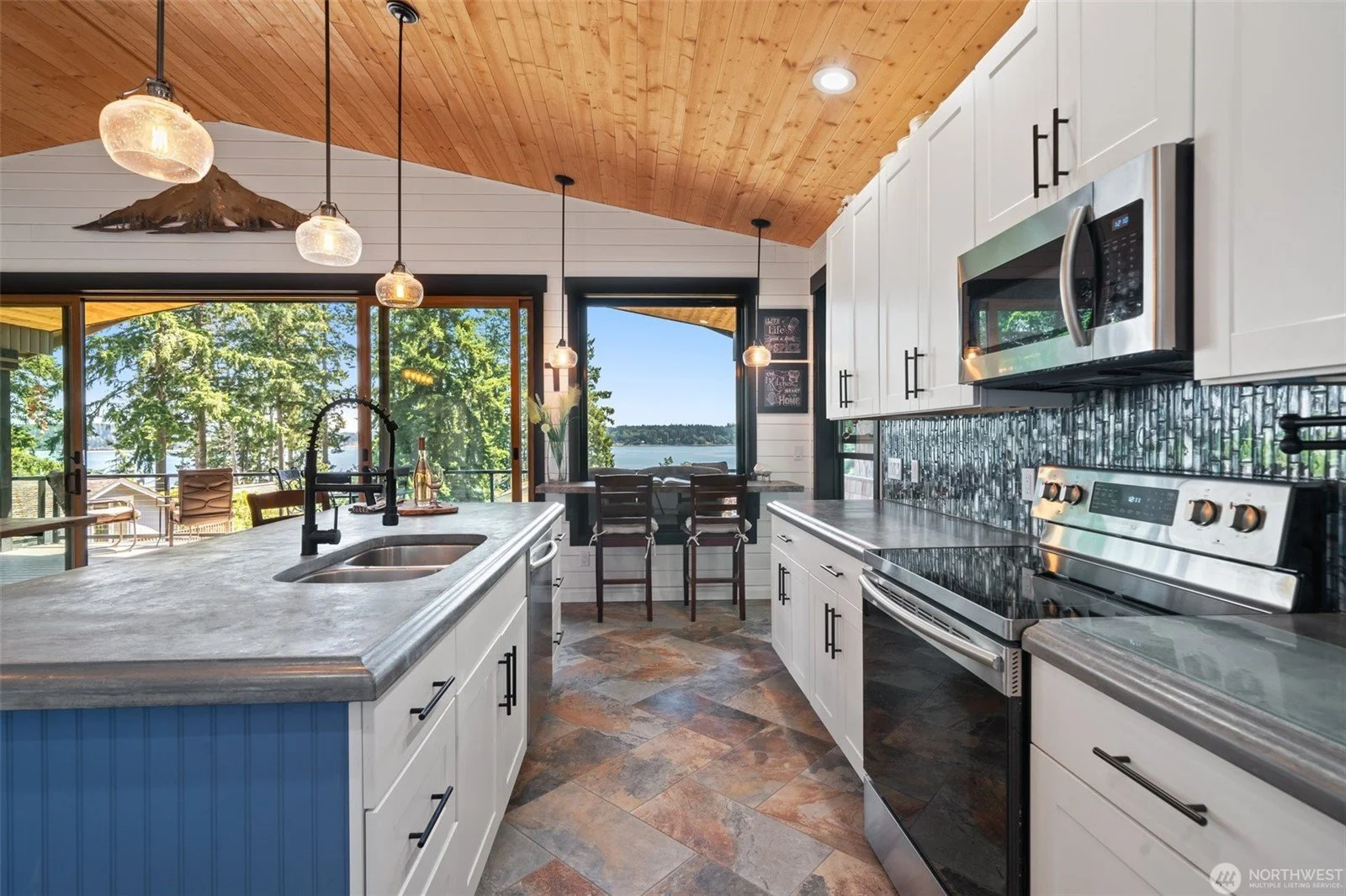 Modern kitchen with white cabinets, black countertops, stainless steel appliances, a colorful tile backsplash, and a view of water and trees through large windows. Pendant lights hang over a kitchen island with a double sink.