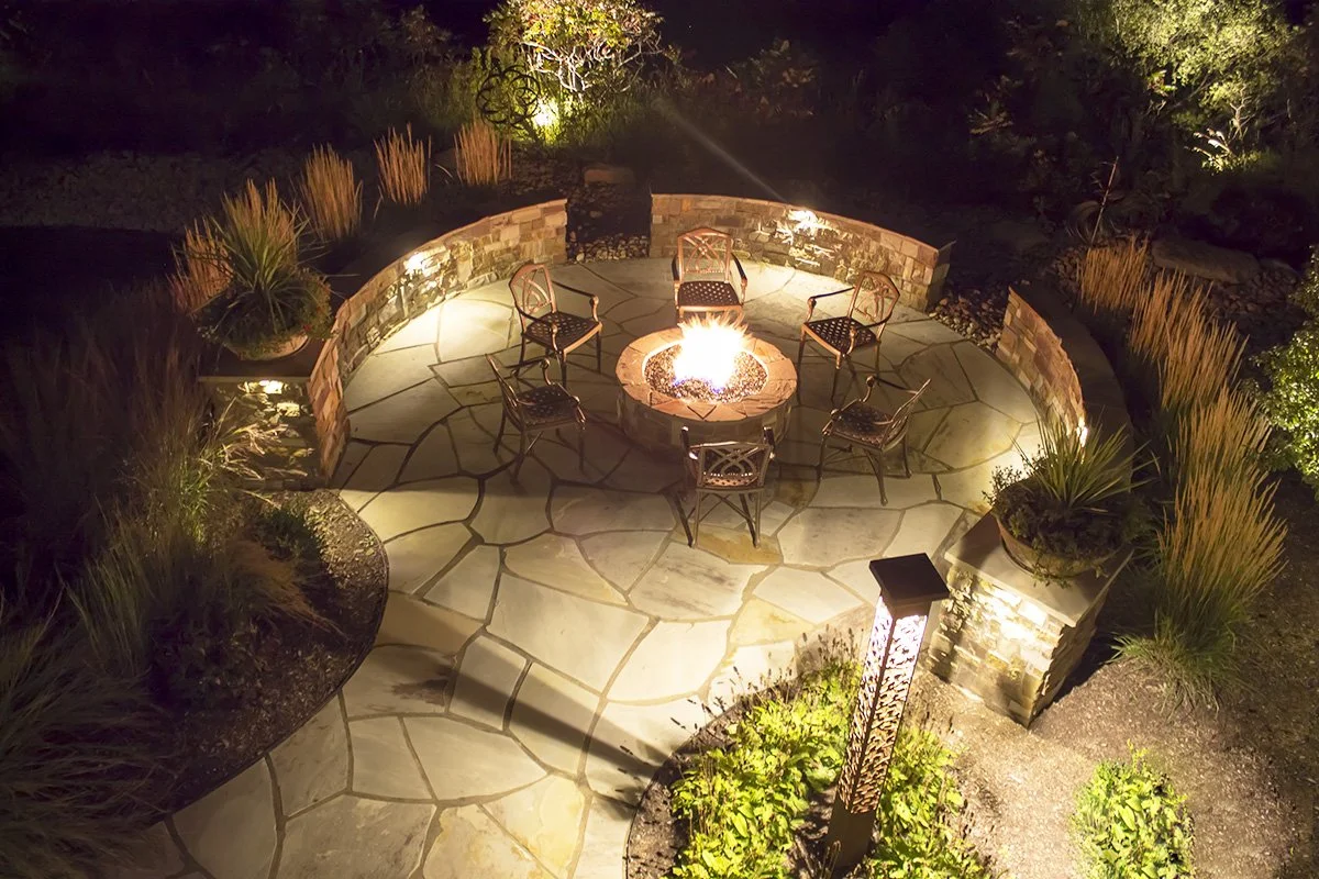 An outdoor fire pit surrounded by six chairs on a circular stone patio at night. Decorative landscaping with plants and shrubs is illuminated by pathway lighting.