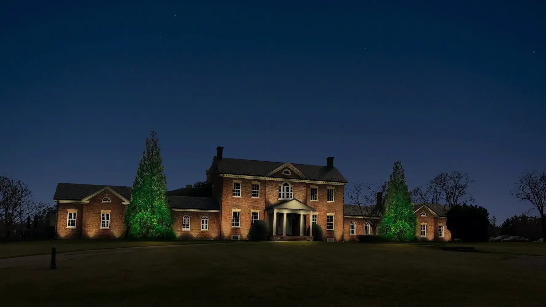 A custom lighting rendering showing a nighttime view of a large brick house with illuminated trees in front and a dark sky with some stars.