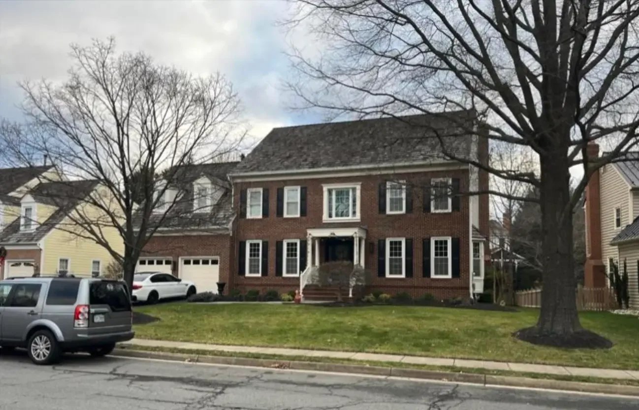 A large brick house with black shutters and white trim, surrounded by a lawn and large trees, with cars parked in the driveway and on the street.