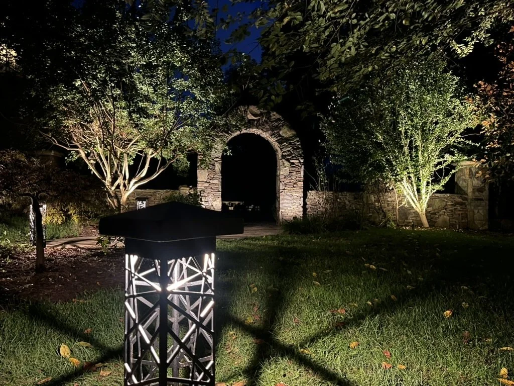 Nighttime garden scene with lit pathway, trees, and stone archway illuminated by lights.