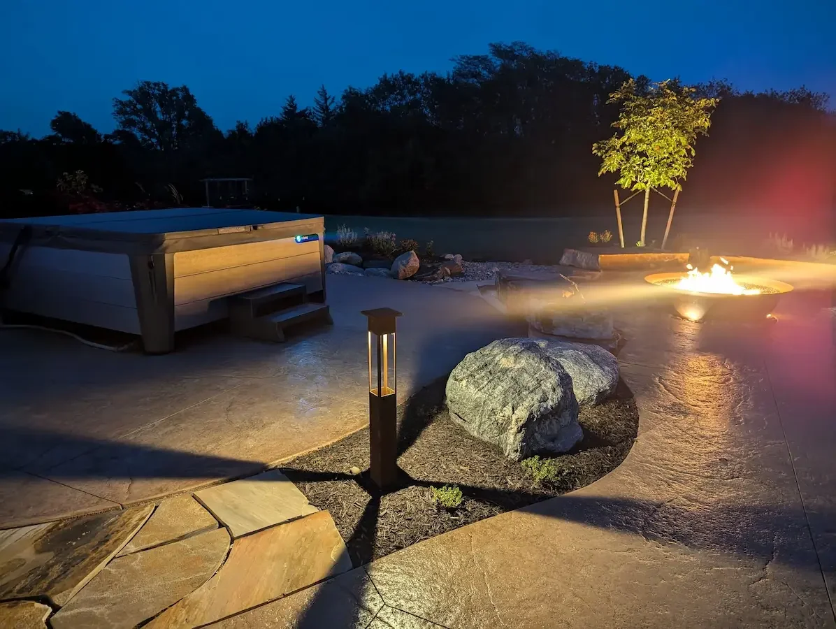 Nighttime scene of an outdoor patio with a hot tub, a lit fire pit, and a small tree illuminated by a nearby lamp. Rocks and trees are in the background.