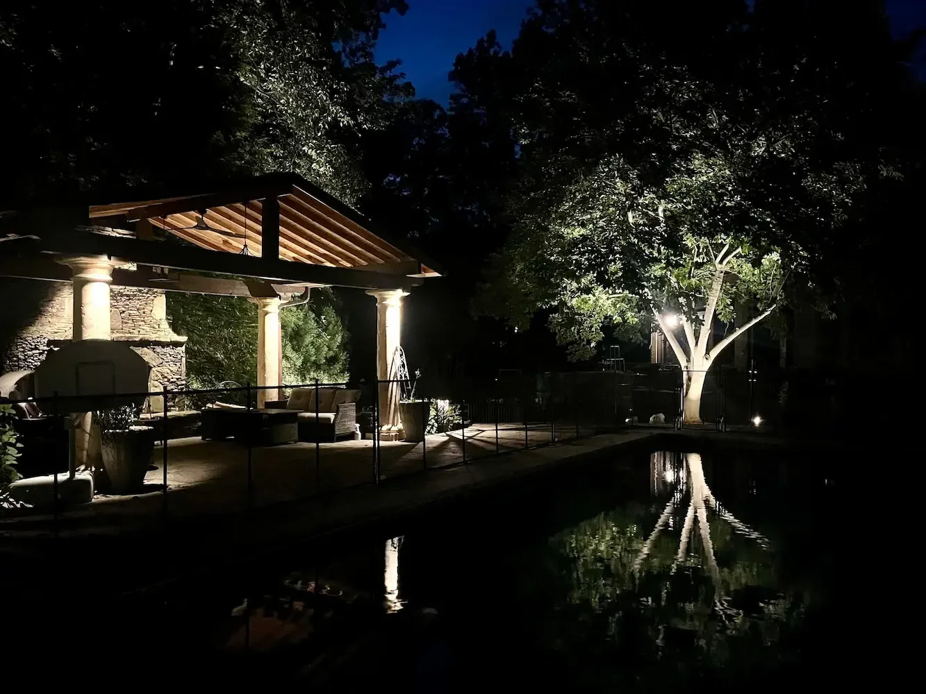 A lit outdoor seating area beside a swimming pool at night, with a tree and a gazebo, illuminated by soft lighting.