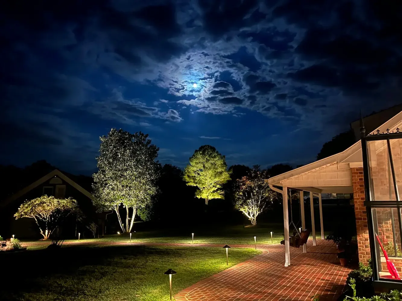 Nighttime scene featuring a house with a brick patio, illuminated trees, and a partly cloudy sky with the moon shining behind clouds.