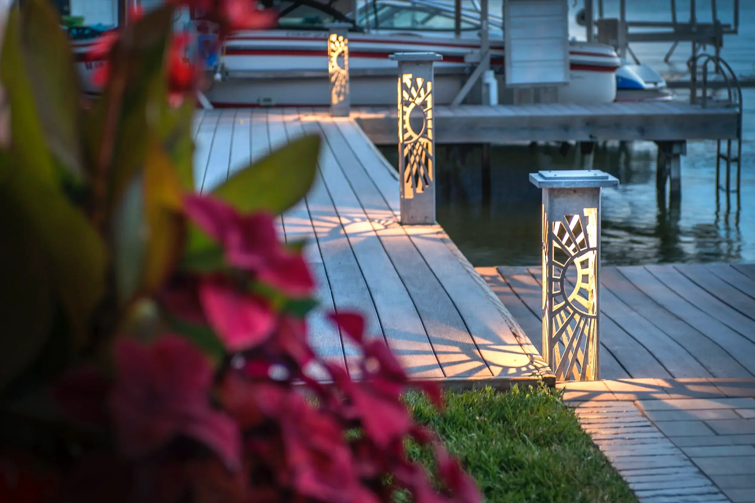 A wooden dock beside a body of water with three decorative lanterns illuminated along the dock. A boat is docked at the end of the dock. Pink flowers and green leaves are in the foreground.