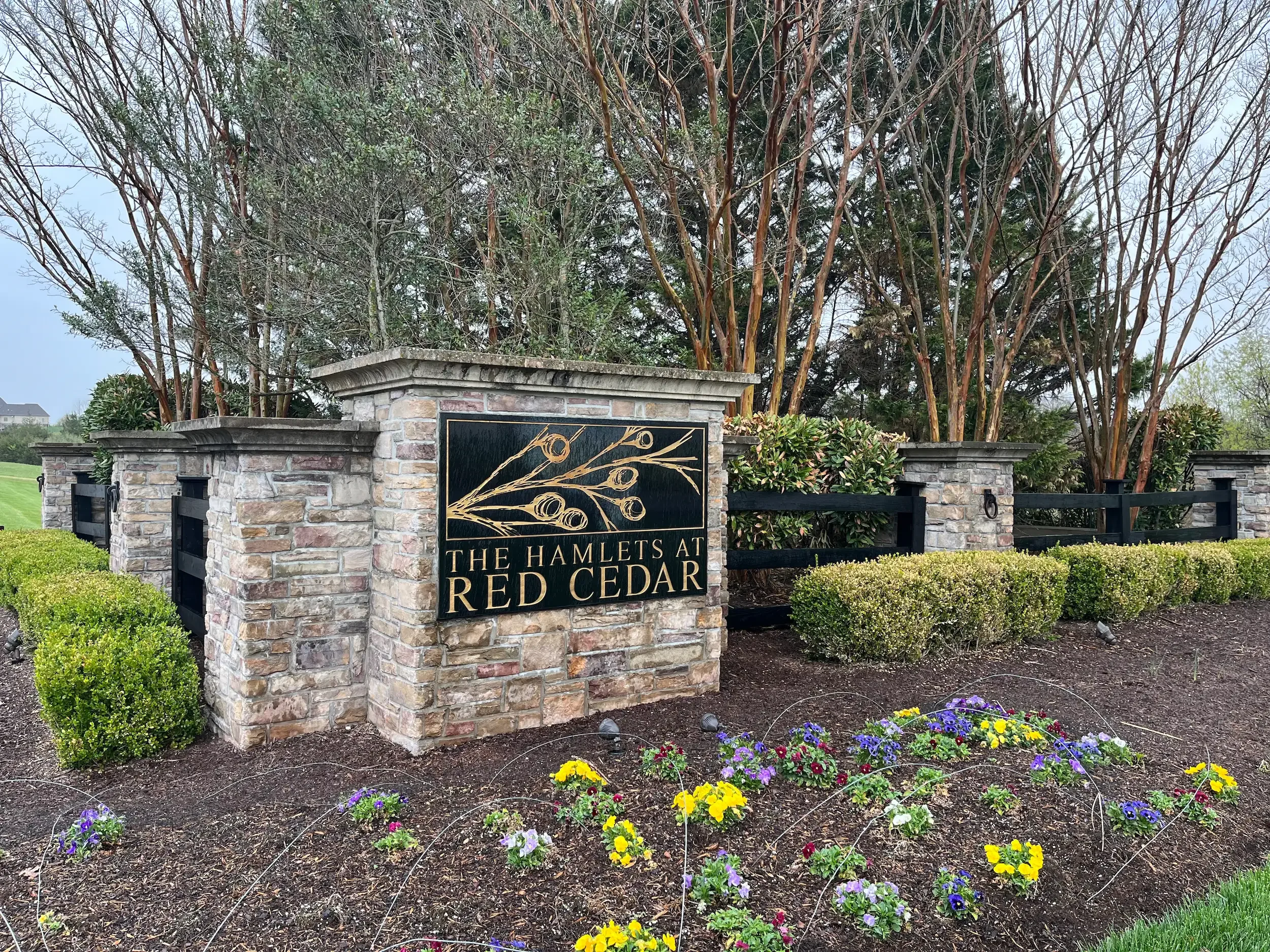 A stone wall with a black and gold sign reading 'The Hamlets at Red Cedar' in front of trees and colorful flower bed.