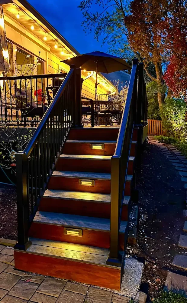 Outdoor wooden staircase with added yellow lights leading to a patio in the evening, surrounded by trees with autumn foliage and a house with string lights.