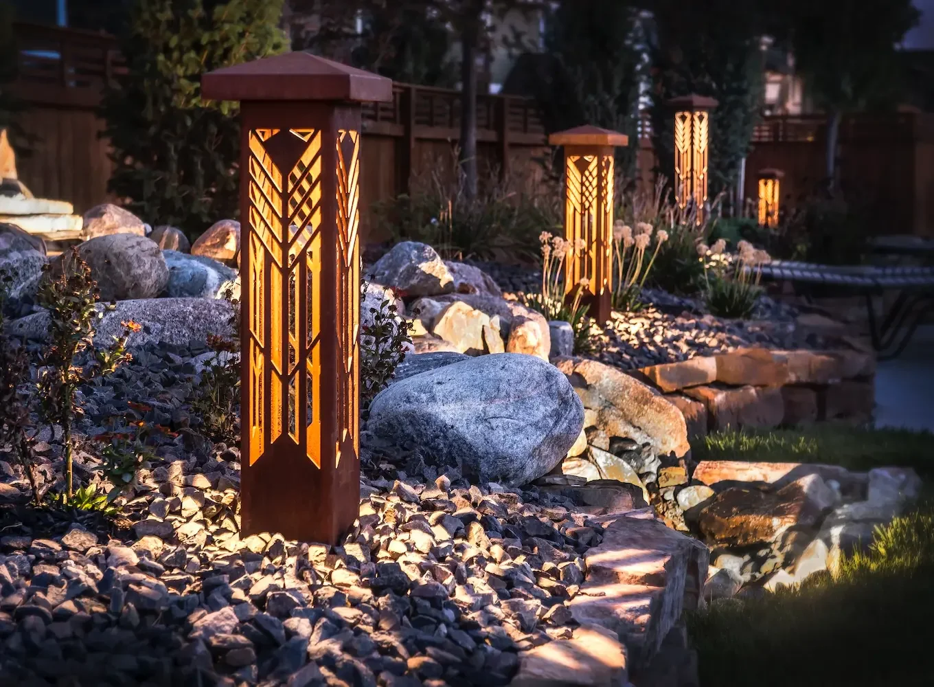 Outdoor garden pathway illuminated by decorative bollard lights with warm light, surrounded by rocks, plants, and flowers at dusk.