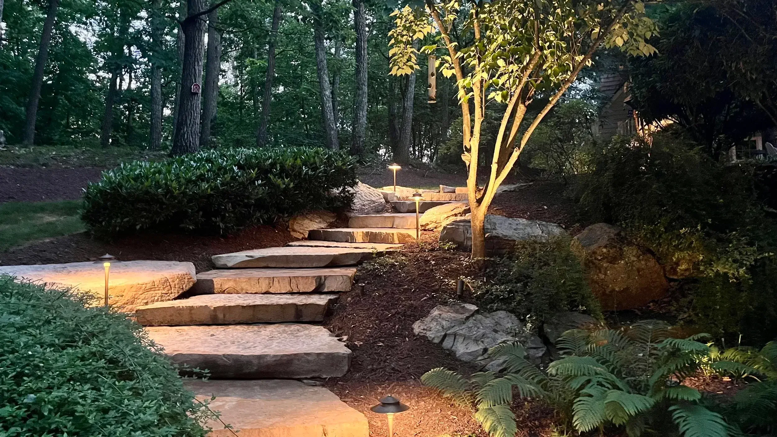 Stone pathway illuminated by small garden lights winding through a landscaped yard with trees, bushes, and rocks in the evening.