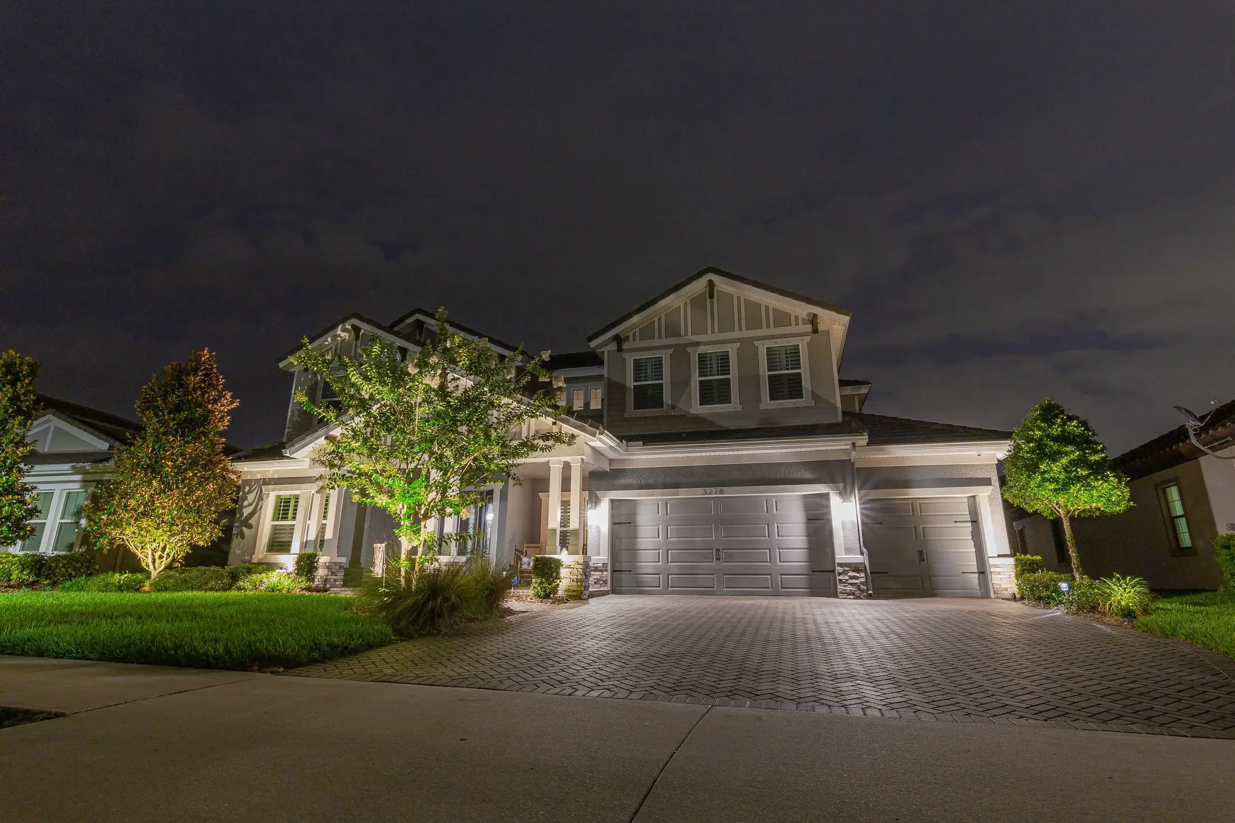 Nighttime exterior view of a modern two-story house with illuminated entrance, garage, landscaped front yard with trees, shrubs, and grass, and cloudy sky in the background.