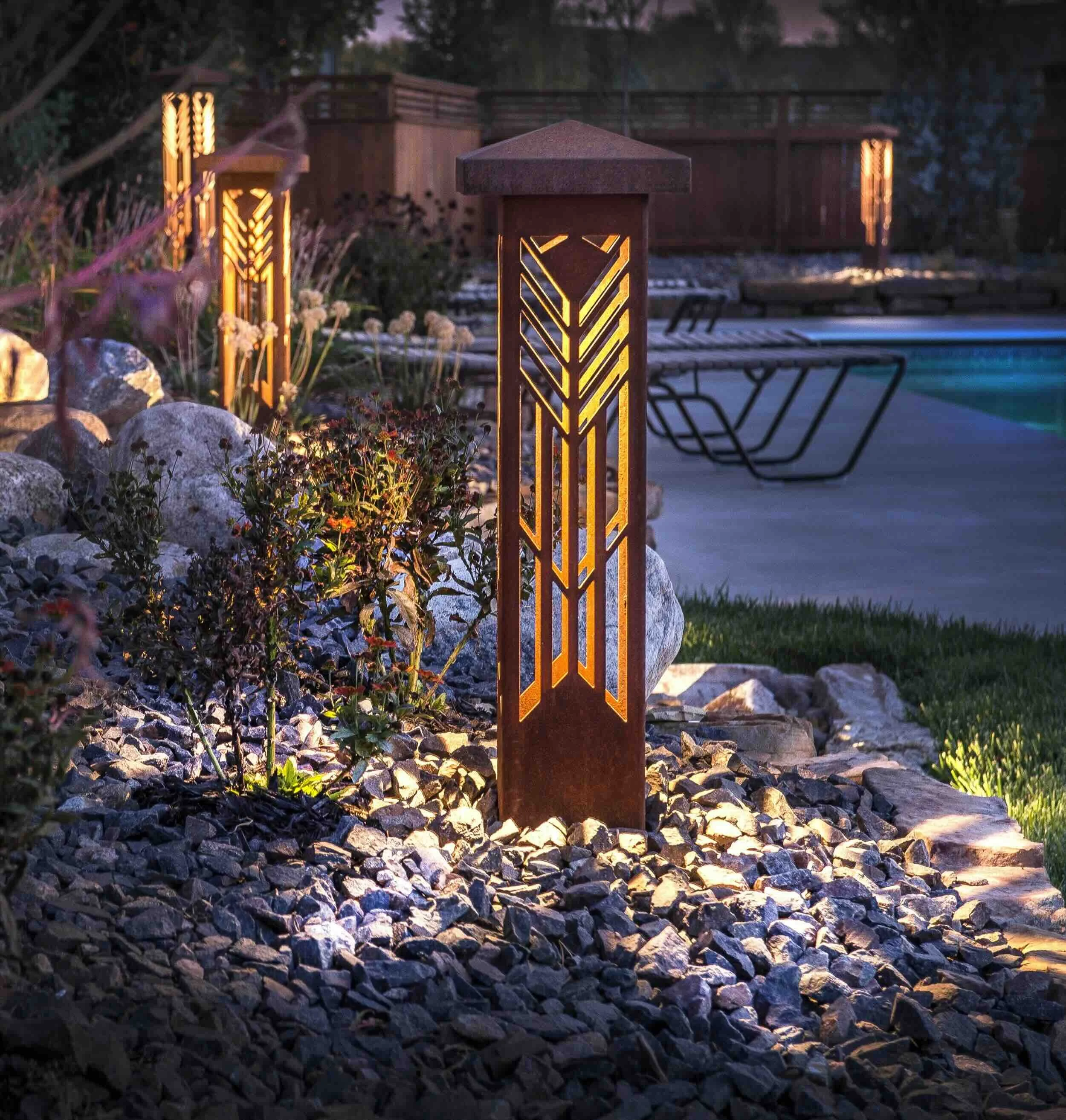 Outdoor garden with decorative pathway lighting fixtures, pebbles, plants, and a pool area in the background.