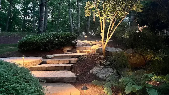 Outdoor garden pathway with stone steps, illuminated by pathway lights, surrounded by trees, bushes, and plants in the evening.