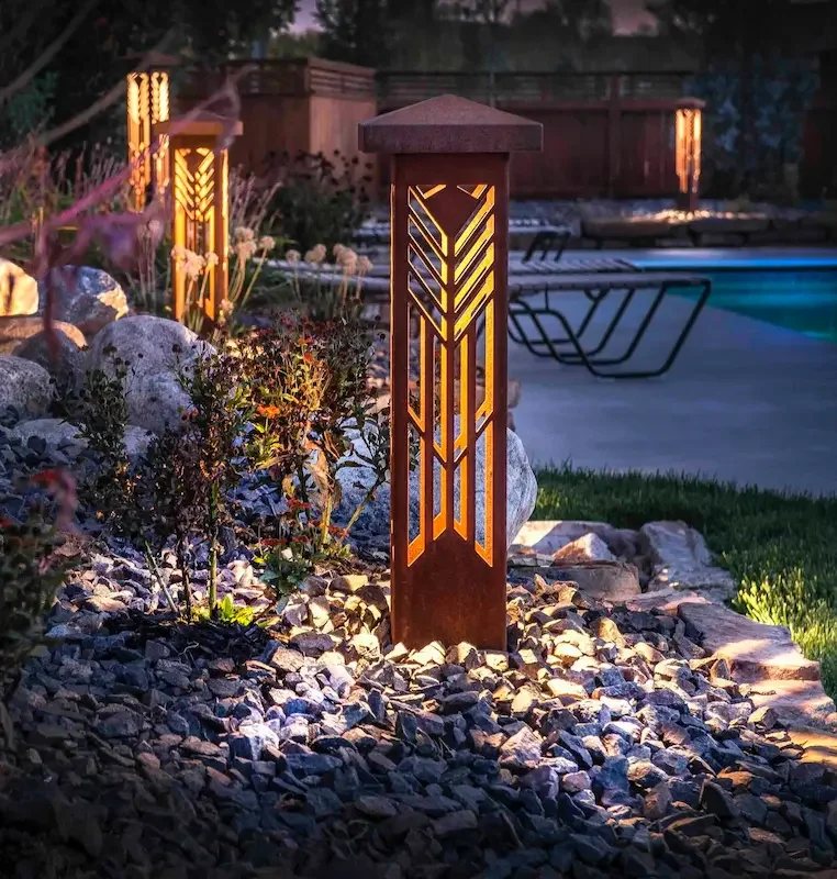 Decorative outdoor garden lamp post with geometric design, illuminated, surrounded by rocks and plants near a swimming pool at dusk.