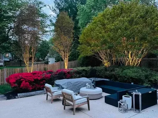 A backyard patio area with two white chairs, a black sectional sofa, and a white ottoman, surrounded by shrubs, flowering plants, and illuminated trees near a wooden fence at dusk.
