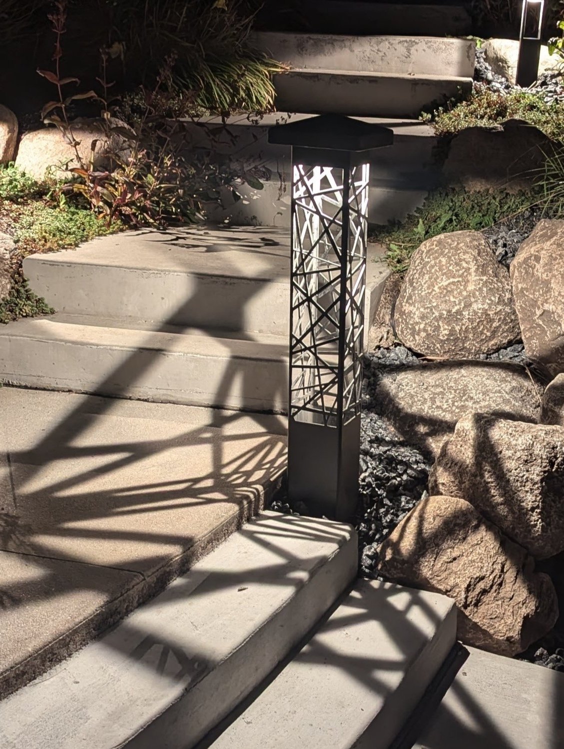 Outdoor pathway with steps, decorative metal lamp casting shadows, surrounded by rocks and plants.
