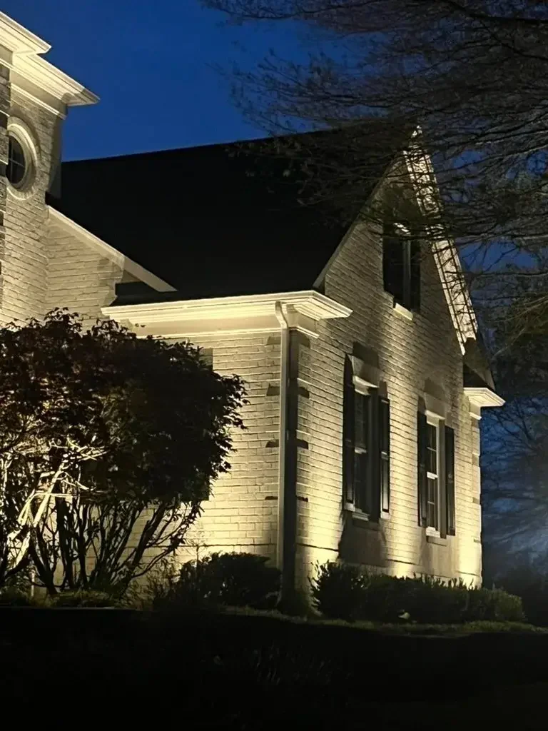 A historic house illuminated by outdoor lighting at night, showing brick walls, multiple windows with decorative shutters, and a gabled roof. There are bushes and a tree in front of the house.