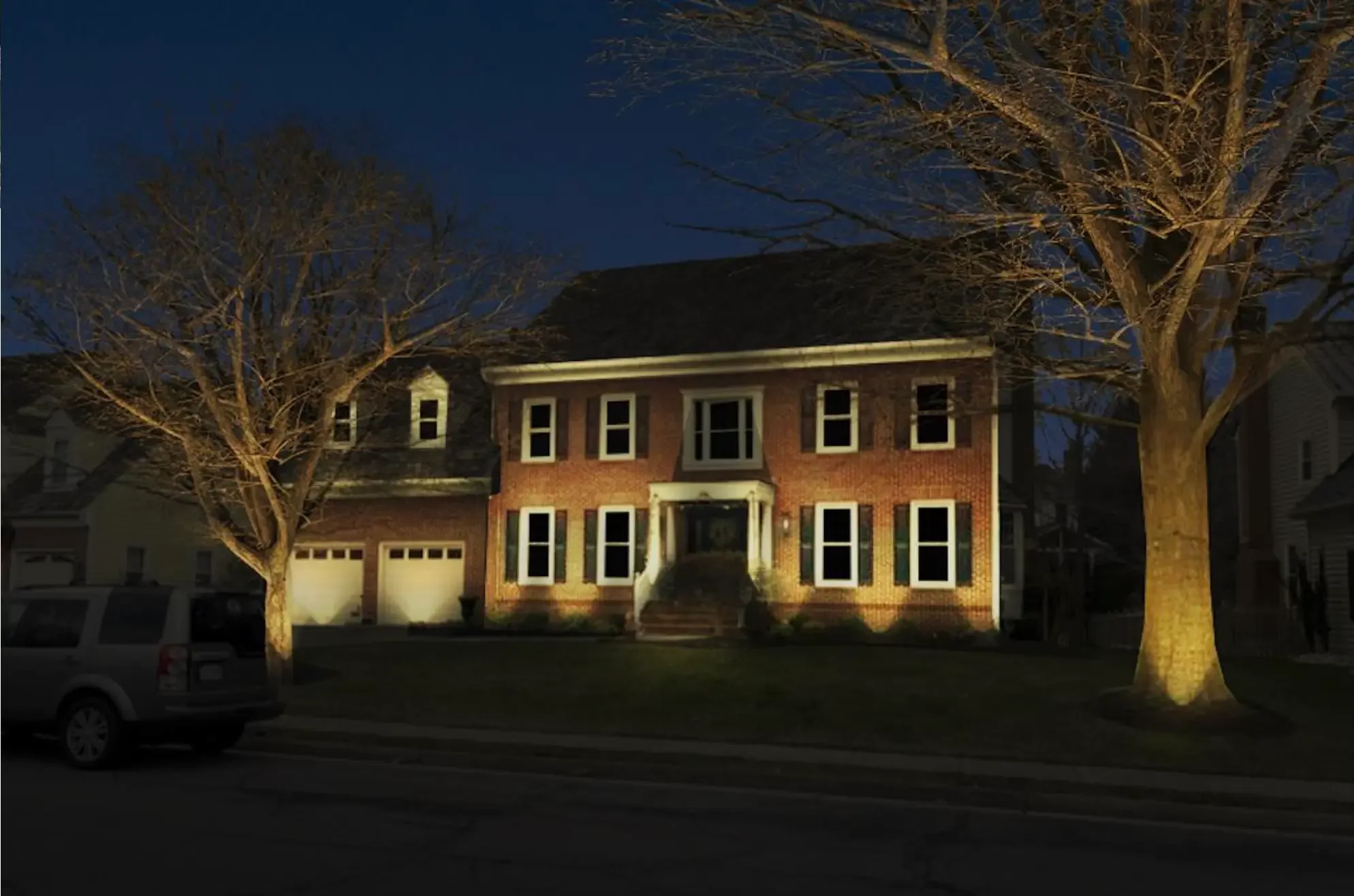 A large brick house at night with exterior lights illuminating the front facade, two large leafless trees flanking the house, and a parked SUV on the street in front.