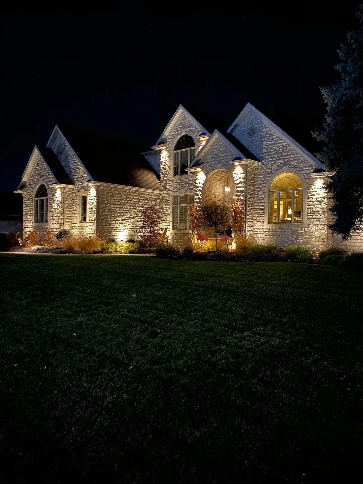 Illuminated large stone house at night with garden and trees in front.
