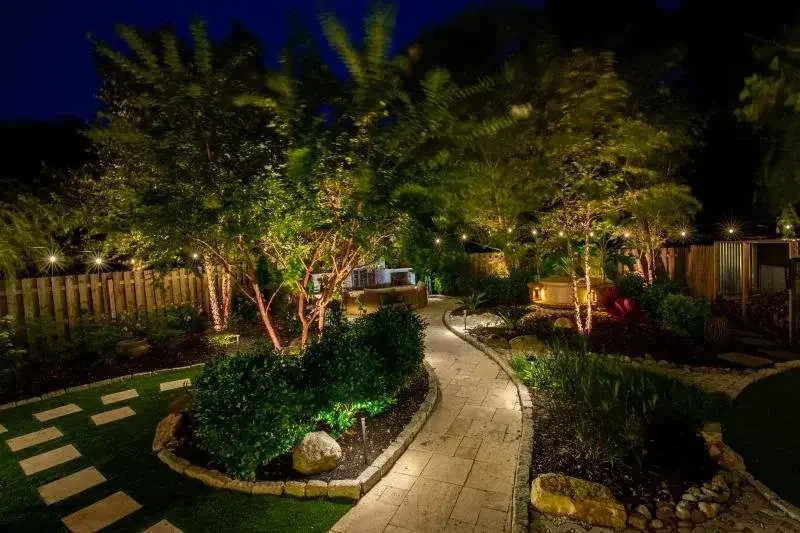 Nighttime garden with illuminated trees, a paved winding pathway, shrubs, rocks, and a wooden fence in the background.