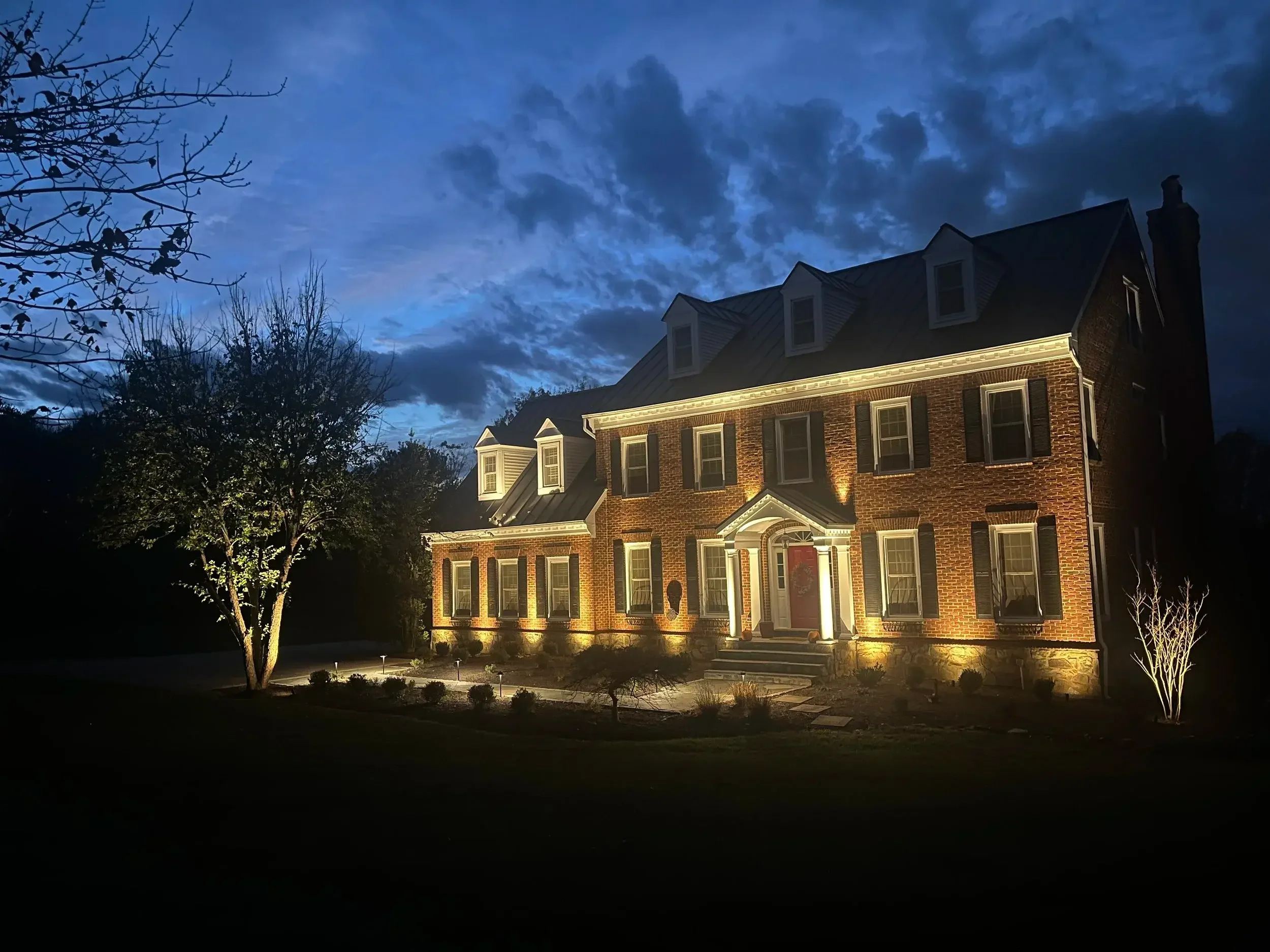 The warm glow of uplighting highlights the timeless elegance of this Colonial home as the evening sets in.