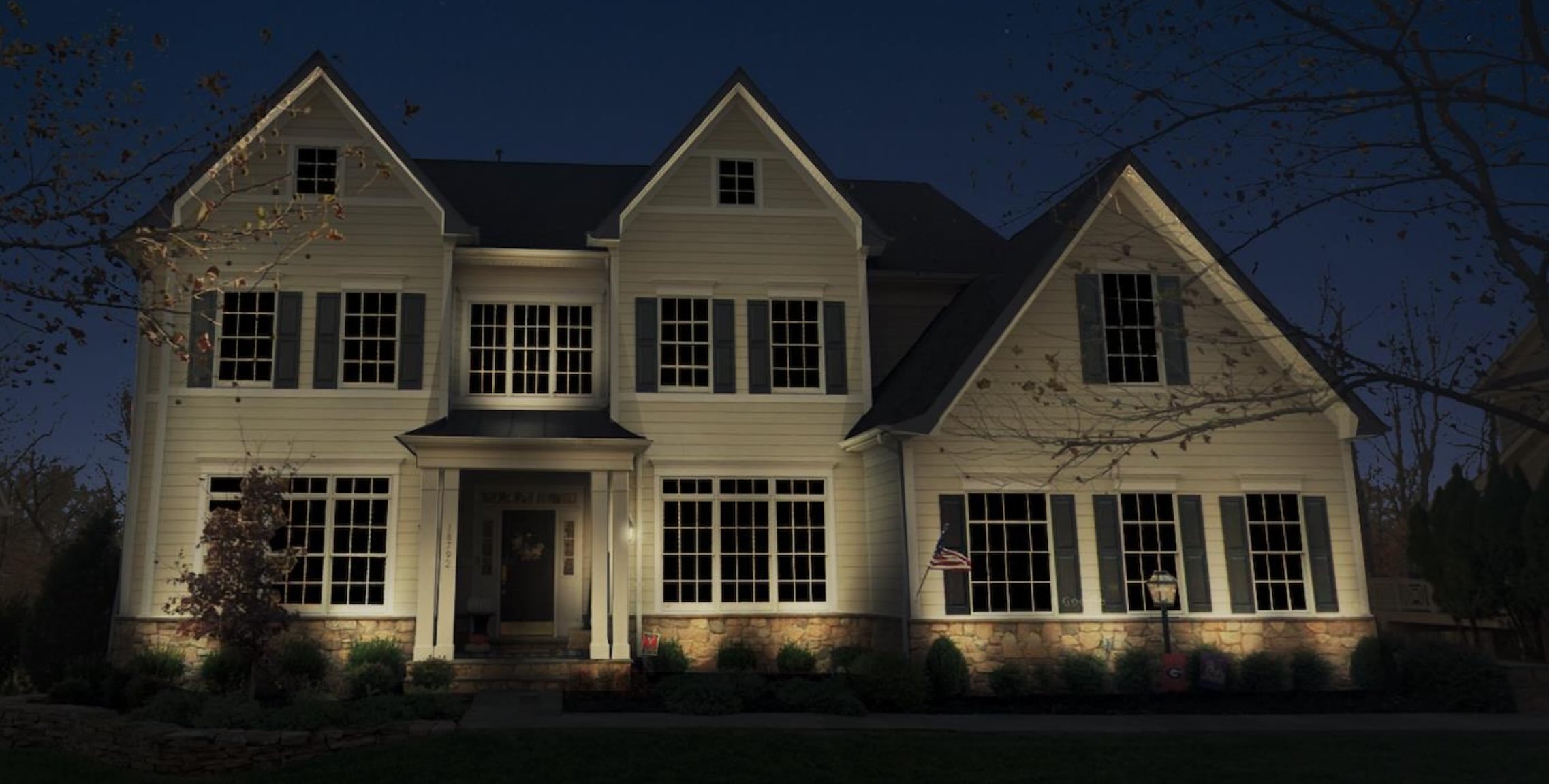 Large two-story house with illuminated lights at night, featuring white siding, numerous windows, and a stone base, surrounded by trees.