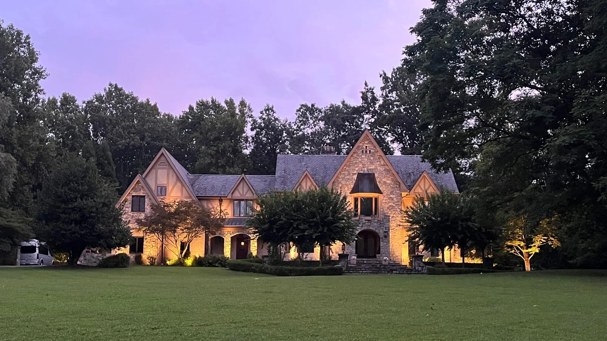 A large, stone and wood house with a steep roof, illuminated from below, surrounded by trees and a well-kept lawn during dusk.