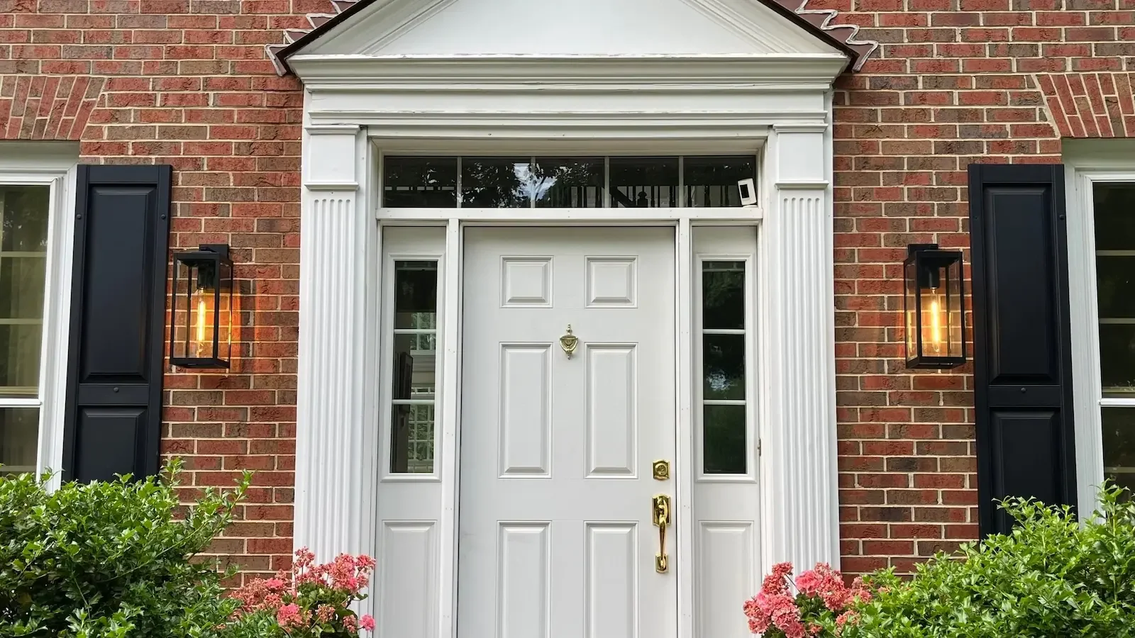 White front door with brass hardware, flanked by modern sidelights and topped with transom window, set in a red brick house.