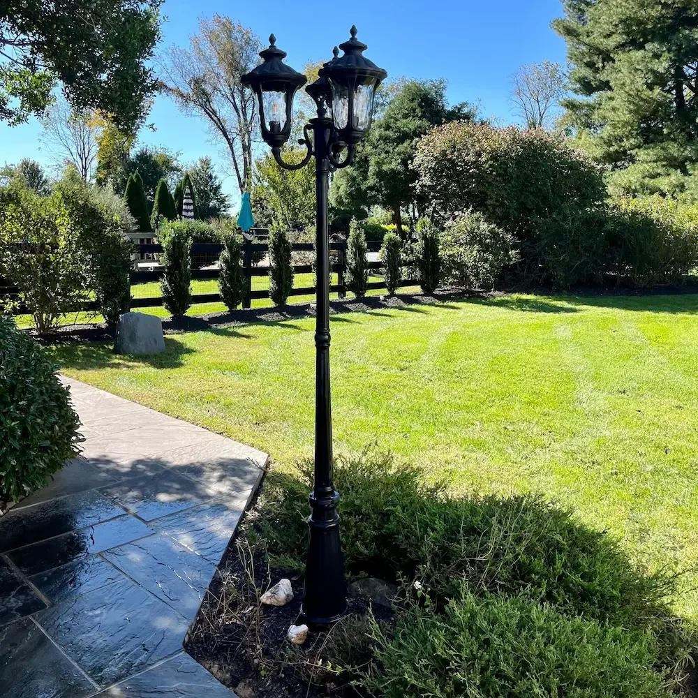 A black three-lantern street lamp in a grassy backyard garden, surrounded by bushes and trees, with a stone pathway on the left and a wooden fence in the background.