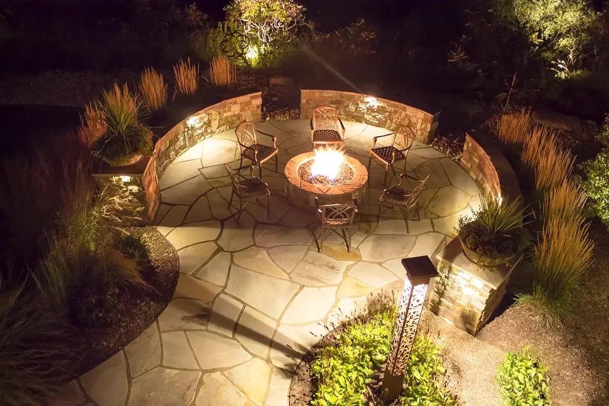 Night view of an outdoor fire pit patio with six chairs arranged around it, surrounded by landscaped plants and tall grasses, illuminated by outdoor lighting.