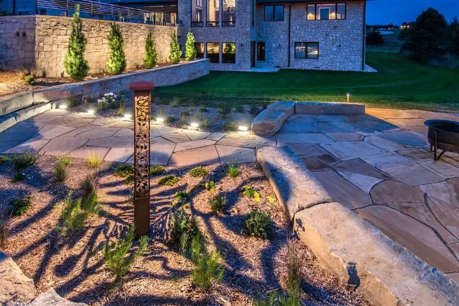 A backyard patio area at dusk with landscaped garden beds, small shrubs, a firepit, and decorative lighting, with a stone house in the background.